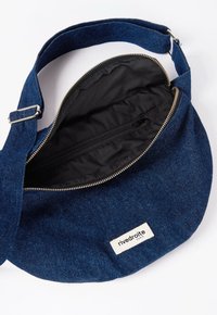 Sac en denim rond avec un extérieur bleu, un intérieur noir et une fermeture éclair. Il dispose d'une bandoulière et d'une étiquette avec le logo "rivedroite".