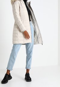 Ljusbeige puffjacka med avtagbar huvtröja i päls, med dragkedja och horisontell quiltad mönster. Matchas med blå jeans och svarta sneakers.