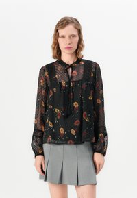 Blusa nera trasparente con stampa floreale, texture a pois e scollatura rotonda. Caratterizzata da maniche lunghe e un dettaglio annodato al collo.