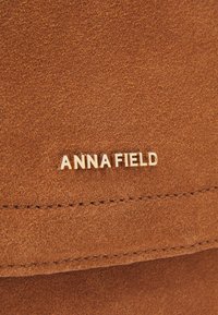 Ruskea nubukkinakangas, jossa on sileä pinta. Kulta "ANNA FIELD" -logo, joka korostaa. Näkyvä ompelu lisäominaisuutena parantaa kokonaismuotoilua.