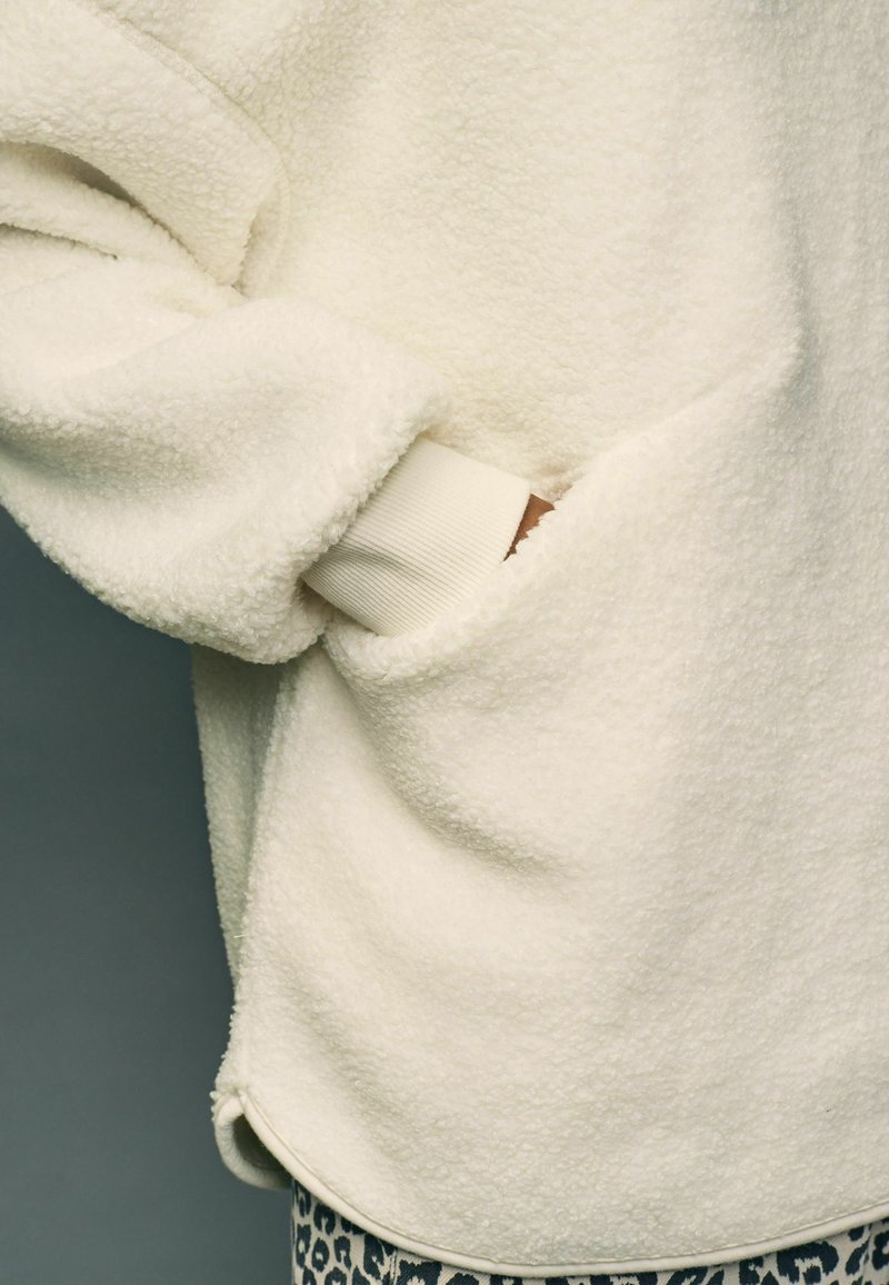 Personne portant une veste polaire couleur crème avec la main dans la poche latérale, montrant un bord-côte et un pantalon à imprimé léopard.