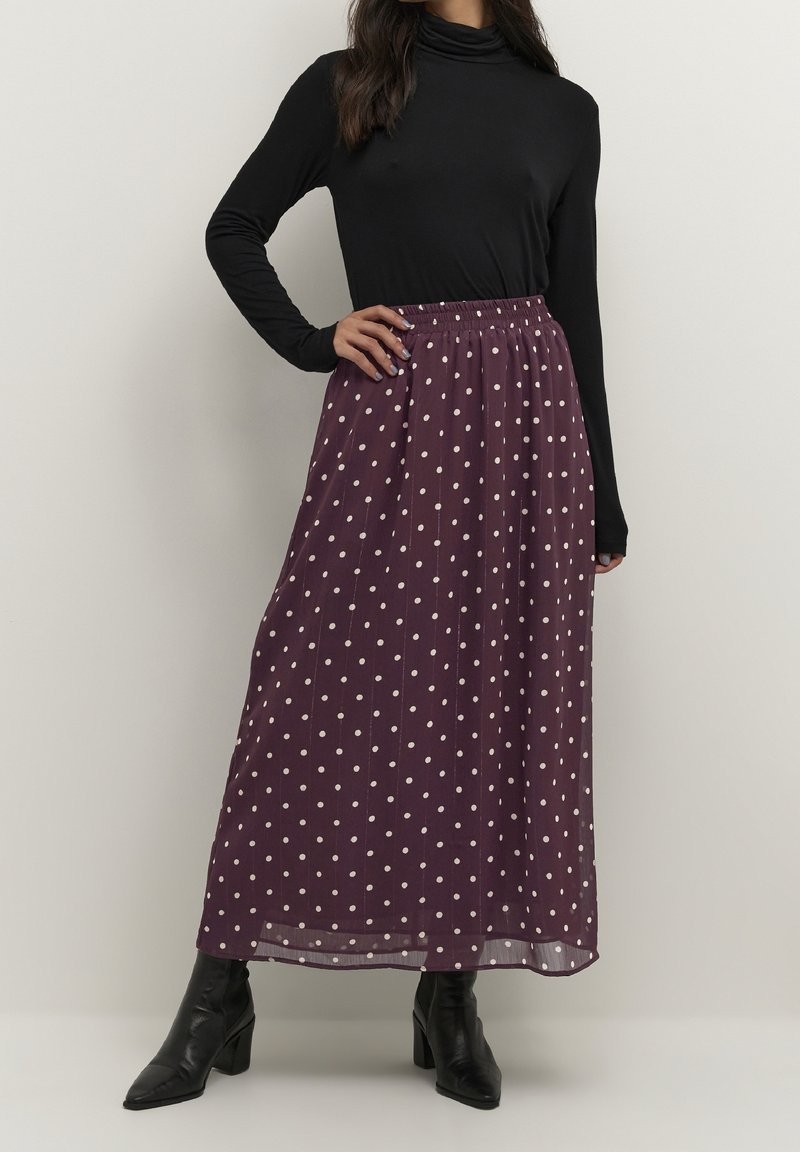 Femme portant un haut noir à manches longues, une jupe longue violette à pois blancs et des bottines noires, debout la main sur la hanche.
