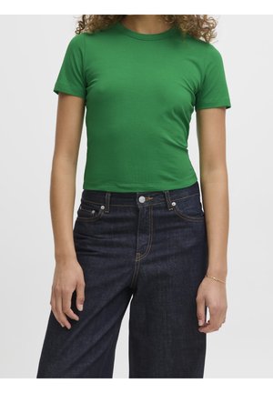 Osoba ubrana w dopasowaną zieloną koszulkę z krótkim rękawem oraz wysokie, szerokie granatowe spodnie dżinsowe na tle jednolitego tła.