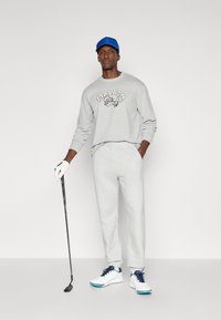 Γκρι φούτερ με γραφικό "MALBON", αντίστοιχες γκρι φόρμες, μπλε καπέλο, λευκά παπούτσια γκολφ με μπλε λεπτομέρειες και σκεπάρνι στο χέρι.