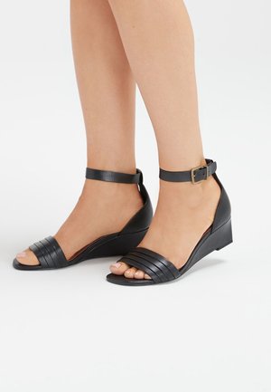 Jambes portant des sandales compensées noires avec des brides à la cheville et des bandes avant texturées sur fond blanc.