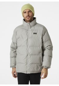 Chaqueta acolchada gris claro con cuello alto, costuras visibles y logotipo "HH" en el pecho. Lleva puesta una gorra amarilla clara de textura.
