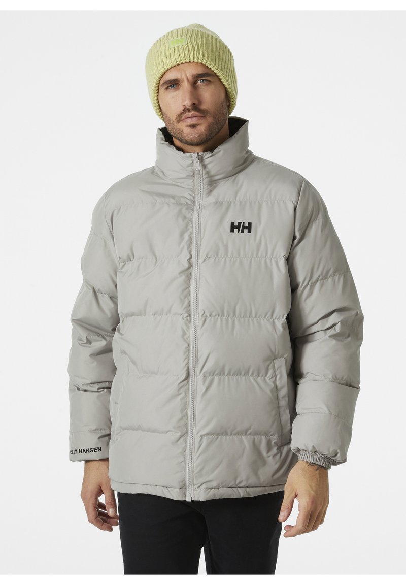 Chaqueta acolchada gris claro con cuello alto, costuras visibles y logotipo "HH" en el pecho. Lleva puesta una gorra amarilla clara de textura.