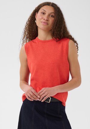 Vrouw met lang krullend haar draagt een rood mouwloos topje en een donkere denim rok, staat met de handen samengevouwen ter hoogte van de taille tegen een effen achtergrond.