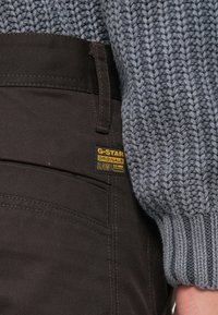 Temno rjave jeans hlače iz denima z pletenim sivim puloverjem, ki ima vidno oznako G-Star Originals na boku.