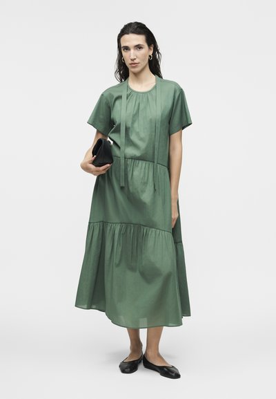 Γυναίκα που φοράει πράσινο φόρεμα μέσης μήκους με στρώσεις και κοντά μανίκια, κρατώντας μαύρο τσαντάκι και φοράει μαύρες μπαλαρίνες πάνω σε λευκό φόντο.