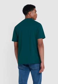 Uomo di spalle che indossa una t-shirt a maniche corte verde scuro e jeans blu su uno sfondo chiaro.