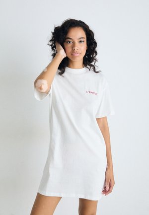 Kobieta z kręconymi włosami, nosząca białą sukienkę T-shirt z napisem "L'AMOUR", dotykająca swoich włosów, stojąca na tle jednolitego, białego tła.