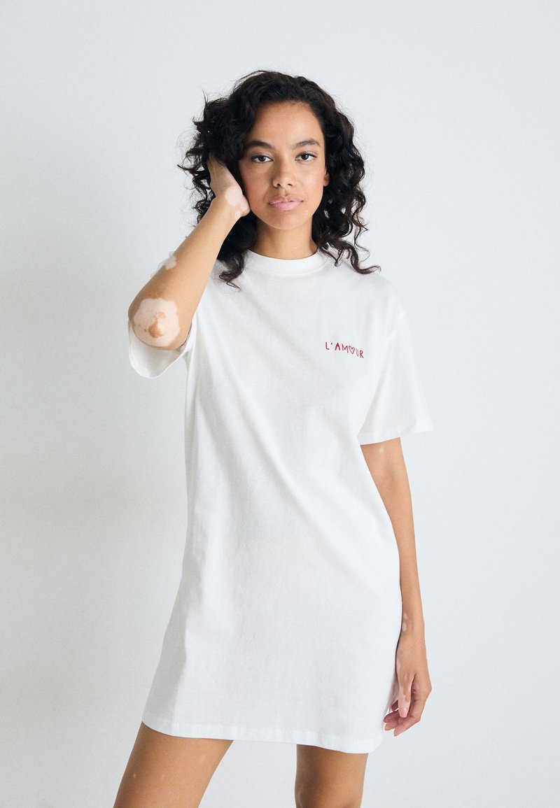 Donna con capelli ricci che indossa un vestito t-shirt bianco con la scritta "L'AMOUR", mentre tocca i suoi capelli, in piedi di fronte a uno sfondo bianco semplice.