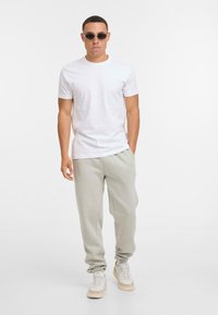 Man in ronde zonnebril, witte korte mouwen t-shirt, lichtgrijze trainingsbroek en witte sneakers staat tegen een eenvoudige witte achtergrond.