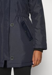 Manteau d'hiver bleu marine foncé avec taille élastique, poche à rabat boutonnée et boutons noirs, porté avec un pantalon noir et une main visible sur le côté.