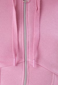 Sudadera con cremallera en color rosa, que presenta una cremallera plateada, un cordón texturizado con puntas a juego y un acabado de tela suave.