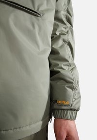 Khaki grön jacka av nylon med en slät textur. Har samlade ärmslut, en ficka med dragkedja och en "CORDURA"-etikett på ärmen.