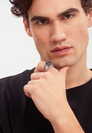 Junger Mann mit dunklem lockigem Haar, der ein schwarzes Hemd trägt und sein Kinn auf seine Faust legt, die mit einem silbernen Ring mit Sternmotiv verziert ist.