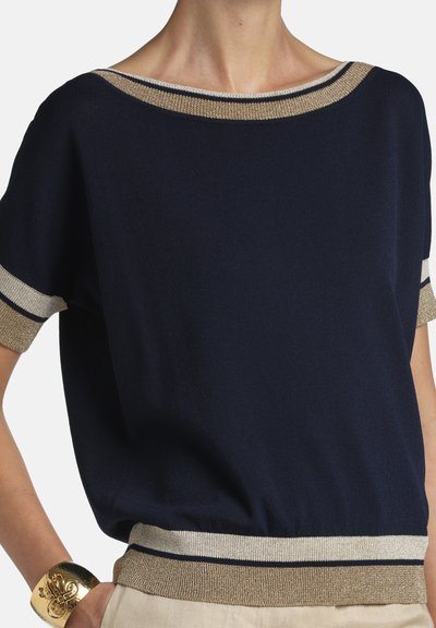 Marineblaues Kurzarmoberteil mit gold-weißen Besätzen am Ausschnitt, den Bündchen und am Saum, getragen von einer Person mit einem goldenen Armreif.