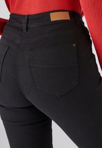 Jeans de denim de tiro alto en color negro, con bolsillos traseros, costuras decorativas y una etiqueta de cuero marrón en la cintura.