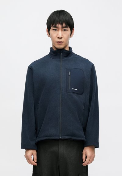 Veste en polaire bleu marine avec col montant, fermeture éclair à l'avant et poche. Présente une texture lisse et un design minimaliste avec un accent de logo.