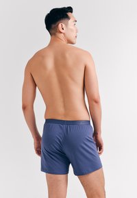 Man met kort zwart haar, gekleed in blauwe boxershorts, staat shirtless met zijn rug naar de camera op een egale lichte achtergrond.