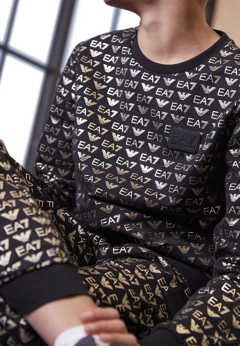 Completo felpa e pantaloni neri con loghi e stemma EA7 dorati. Presenta una scollatura a girocollo e polsini con finitura testurizzata.
