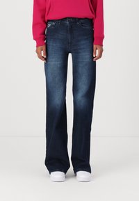 Osoba v tmavě modrých širokých džínách, s dlouhým rukávem v jasně růžové barvě a bílými teniskami, stojící na jednolitém pozadí.