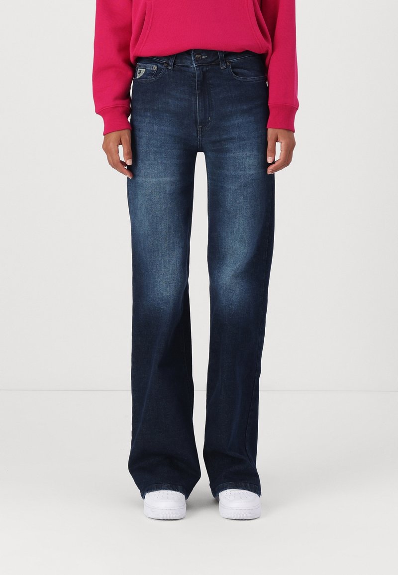 Osoba v tmavě modrých širokých džínách, s dlouhým rukávem v jasně růžové barvě a bílými teniskami, stojící na jednolitém pozadí.