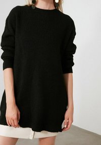 Femme portant un pull noir côtelé oversize à manches longues et de grandes boucles d'oreilles rondes noires et dorées, associé à une jupe courte blanche.