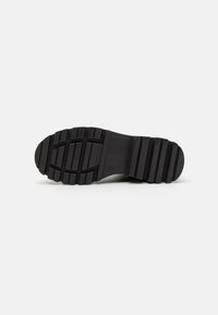 Semelle de chaussure en caoutchouc noir avec un motif de bande de roulement strié et un bout arrondi. Présente des rainures doubles et un logo circulaire près de la base.