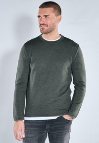 Dunkelgrüner Pullover mit langen Ärmeln und Rundhalsausschnitt aus einem strukturierten Stoff, mit dezenten Nahtdetails und einem darunter getragenen weißen Hemd mit Lagenoptik.