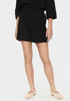 Pantalones cortos de algodón negro con cinturilla elástica, dobladillo doblado y un sutil detalle de costura. Combinados con zapatos de tejido de color claro.