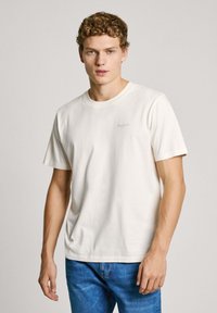 White cotton t-shirt with short sleeves, round neckline, and subtle logo detail on the left chest. Worn with blue denim jeans.