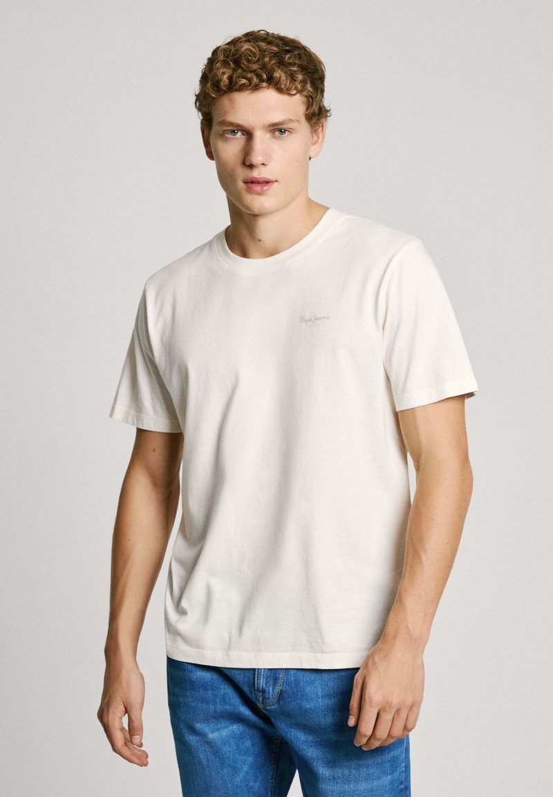 White cotton t-shirt with short sleeves, round neckline, and subtle logo detail on the left chest. Worn with blue denim jeans.