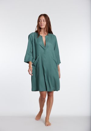 Short, teal dress with a deep V-neck and three-quarter sleeves. Features two side pockets and a relaxed fit, made from smooth fabric.