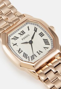 Montre octogonale en or rose avec un cadran blanc texturé, des chiffres romains noirs et un bracelet en acier inoxydable. Logo de marque "FOSSIL" affiché.