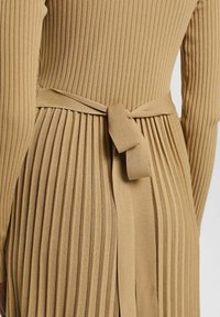 Ribbad, stickad klänning i ljusbrun med en åtsittande överkropp och plisserad kjol. Har ett knytbälte i midjan som är tillverkat av samma material.