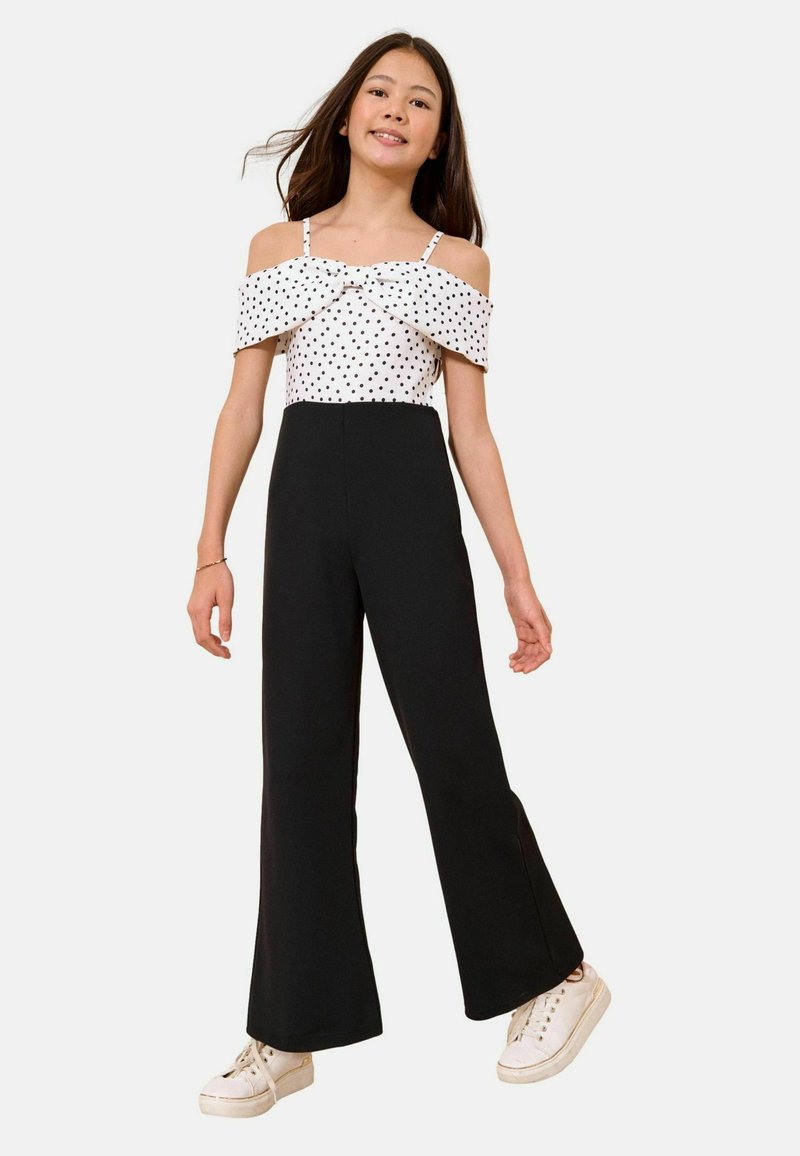 Ragazza che indossa un top bianco a pois con spalle scoperte, pantaloni neri a vita alta e gamba larga e sneakers bianche, in piedi davanti a uno sfondo semplice.