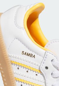 Weiße Ledersneakers mit gelben Akzenten, Zickzackstreifenmuster und goldenem "SAMBA"-Logo. Strukturierte Gummisohle und weiße Schnürsenkel.