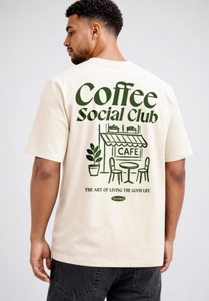 Man wearing cream t-shirt with green "Coffee Social Club" text and cafe illustration on the back, paired with black jeans.