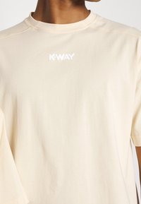 T-shirt di cotone color crema con maniche corte, caratterizzata da un logo bianco in rilievo "K-WAY" sul petto e dettagli di cucitura visibili.