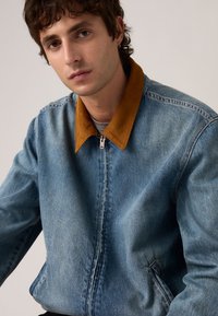 Νεαρός άνδρας με κοντά σγουρά μαλλιά που φοράει μπλε τζιν μπουφάν με καφέ γιακά, καθισμένος μπροστά σε απλό φωτεινό φόντο.