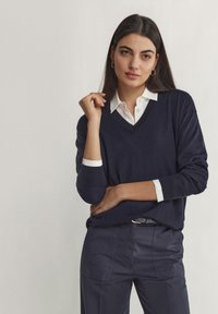 Mörkblå V-ringad tröja i mjukt material, i kombination med en vit skjorta med krage underneath. Modellen står i en avslappnad pose med diskreta accessoarer.