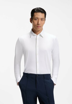 Mann, der ein eng anliegendes weißes Hemd mit langen Ärmeln, Knöpfen und Kragen sowie marineblaue Hose trägt, steht mit den Händen in den Hosentaschen vor weißem Hintergrund.