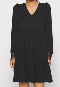 Robe noire en tissu léger, avec un décolleté en V, de longues manches bouffantes avec des accents en dentelle, et un ourlet en volants superposés.