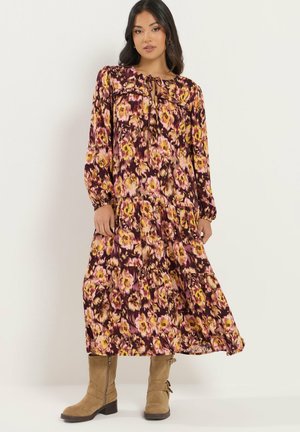 Kvinde iført en langærmet, ankel-lang blomsterkjole i lilla, gul og pink toner kombineret med tan midtkalv støvler stående mod en hvid baggrund.