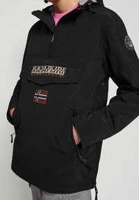 Schwarze leichte Jacke mit einer Fronttasche, die mit dem "Napapijri"-Logo und einem kleinen norwegischen Flaggenpatch versehen ist. Sie verfügt über eine Kapuze und seitliche Reißverschlusstaschen.