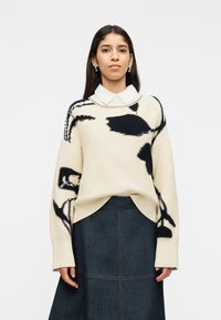 Γυναίκα που φοράει ένα μπεζ πουλόβερ με μαύρα αφηρημένα σχέδια πάνω από ένα λευκό πουκάμισο με γιακά και μια σκοτεινή φούστα από τζιν, στέκεται μπροστά σε ένα λευκό φόντο.