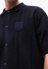 Άνδρας που φοράει μαύρο πλεκτό πόλο με κοντά μανίκια, κάθετες ρίγες και έμβλημα ασπίδας στο αριστερό στήθος, με κουμπιά στο μπροστινό μέρος.